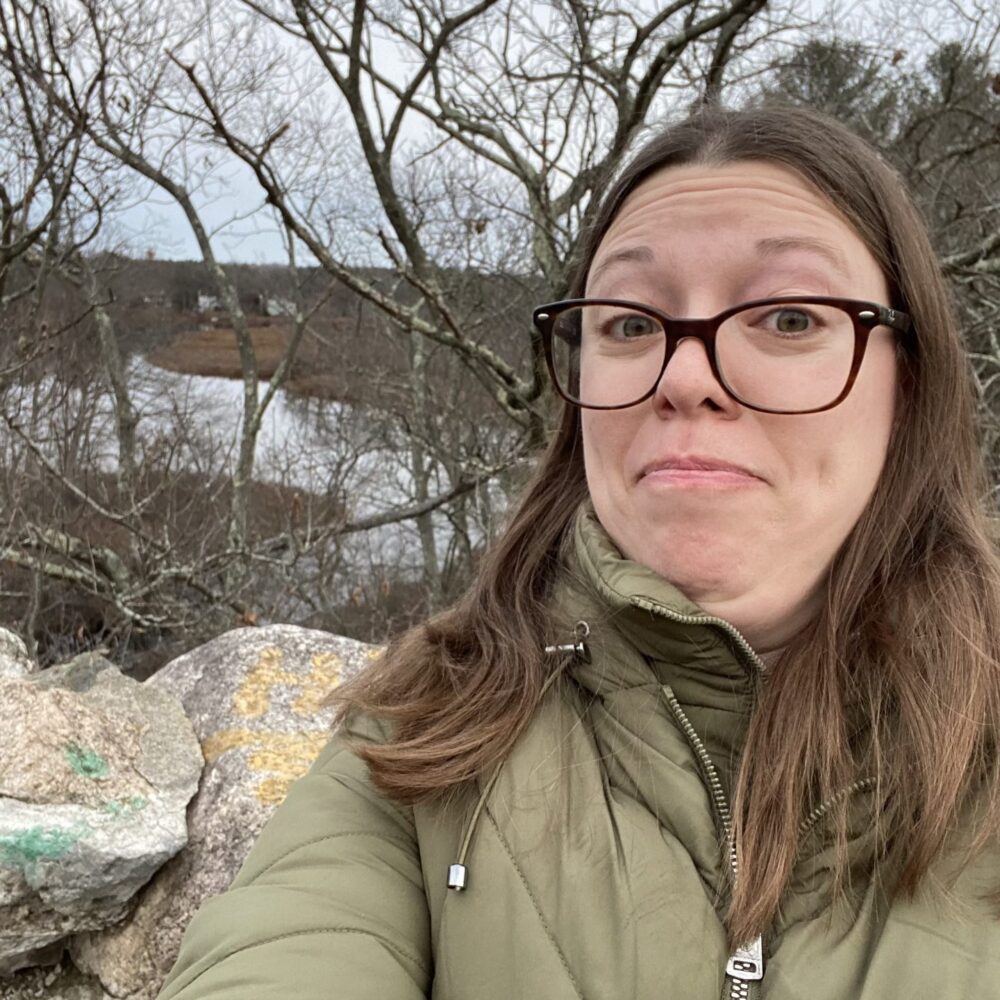 Photo of Amber-Kavka-Warren in a poofy green coat in front of some rocks, barren trees, and water.